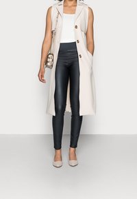 Beige lång väst med öppen front, parat med svarta tajta byxor och ljusbeige klackar. Accessoarer inkluderar en liten väska med ormskinnsmönster.