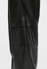 Μαύρο δερμάτινο παντελόνι cargo με υφή. Περιλαμβάνει πλαϊνή τσέπη με καπάκι και κλείσιμο με κουμπί, καθώς και κομψές, λείες ραφές.