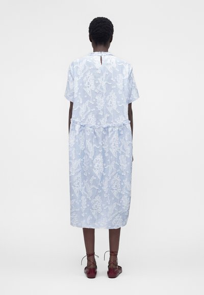 Γυναίκα που φοράει ένα ανοιχτό μπλε midi φόρεμα με λευκή κεντημένη ανθοδέσμη, κοντά μανίκια και μπορντό δαντελωτά παπούτσια, στέκεται με την πλάτη γυρισμένη.