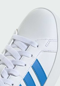 Baskets blanches avec lacets blancs texturés et deux bandes bleues sur le côté, avec des détails cousus sur un matériau en cuir lisse.