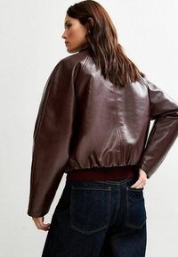 Chaqueta bomber de cuero marrón con dobladillo acanalado, mangas anchas y una textura suave, usada sobre unos jeans de mezclilla oscuros.
