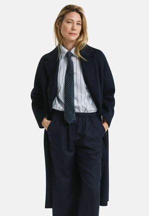 Long navy coat over a light blue striped shirt with a dark tie. Features pleated waistband pants, creating a tailored yet comfortable silhouette.