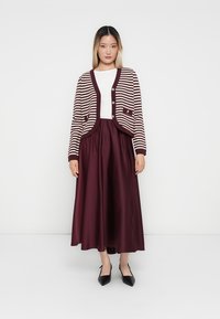 Κόκκινο και κρεμ ριγέ πλεκτό ζακέτα με κουμπιά πάνω από ένα άσπρο πουκάμισο, συνδυασμένο με μία μπορντό σατινέ μακριά φούστα και μαύρες μυτερές γόβες.