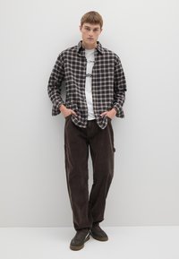 Camicia a quadri in flanella blu scuro e crema, indossata sopra una t-shirt bianca con grafica. Pantaloni di velluto a coste marroni e sneakers scure completano il look.