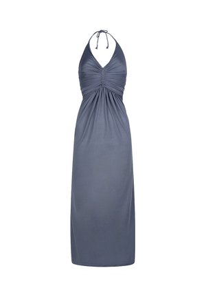 Frau mit blonden Haaren, die ein graues Maxikleid mit Neckholder trägt, steht drinnen neben einer Pflanze und einer cremefarbenen Wand.