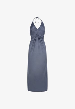 Frau mit blonden Haaren, die ein graues Maxikleid mit Neckholder trägt, steht drinnen neben einer Pflanze und einer cremefarbenen Wand.