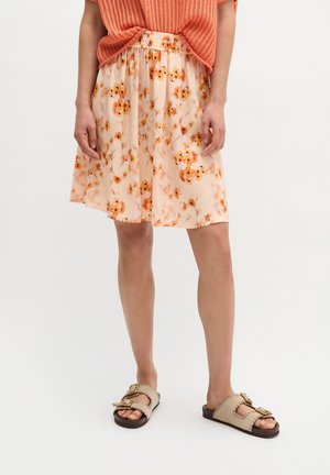 Frau trägt einen knielangen pfirsichfarbenen Blumenrock, ein orangenes Strickoberteil und beige Slip-On-Sandalen mit großen Schnallen und steht vor weißem Hintergrund.