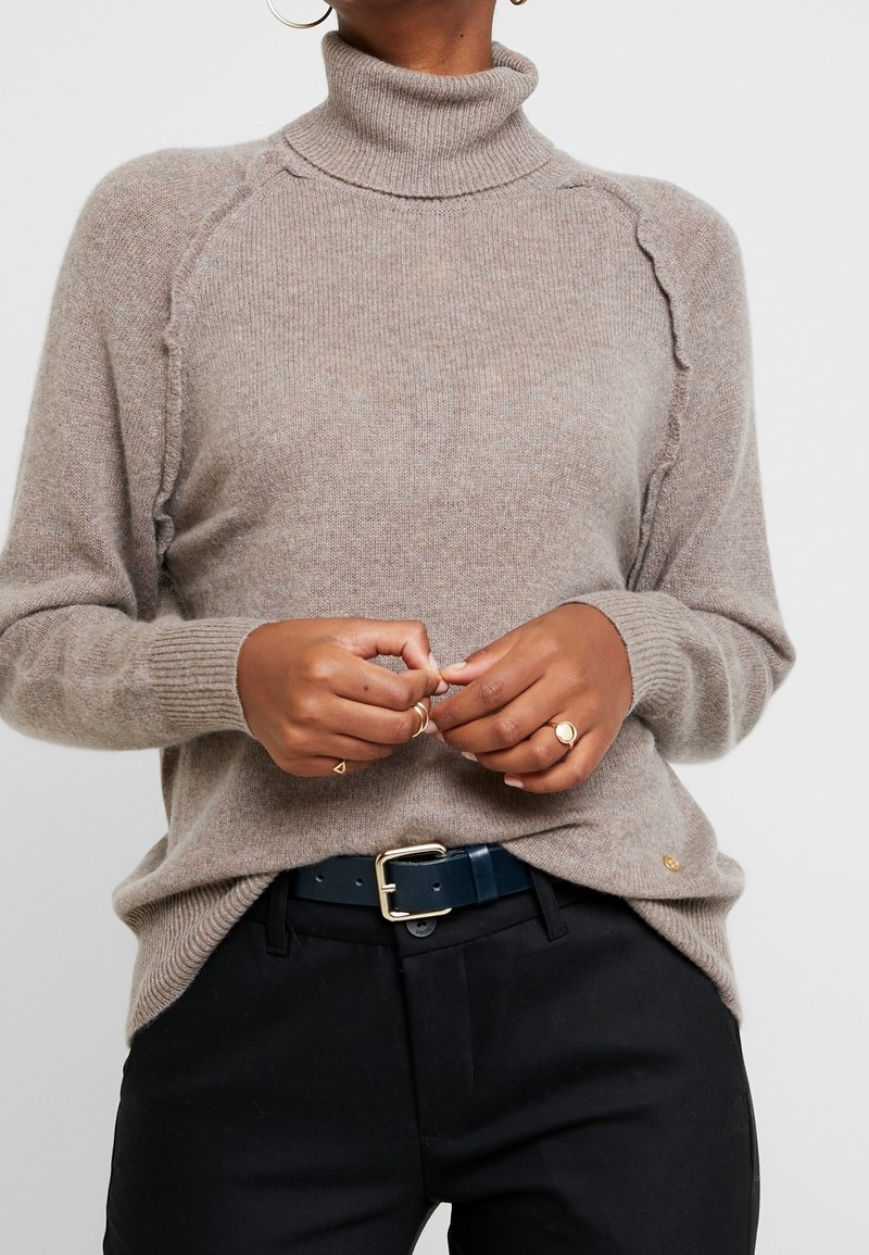 Pull en maille marron clair avec un col roulé haut et des détails festonnés le long des coutures. Porté avec un pantalon noir et accessoirisé avec des bagues.