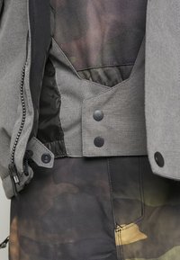 Gros plan sur une veste grise avec une fermeture éclair noire et des boutons pression, portée sur un pantalon à motif camouflage.