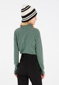 Pull en polaire vert avec col haut, associé à un pantalon de ski noir. Un bonnet en tricot noir et blanc rayé ajoute de la texture et du contraste.