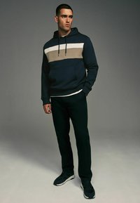 Marineblå hættejakke med en beige og hvid vandret stribe, justerbar snor, frontlomme, parret med sorte bukser og mørke sneakers.