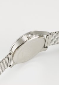 Reloj de acero inoxidable plateado con banda de malla, caja redonda y corona lateral. Superficie lisa y pulida con un diseño limpio y minimalista.