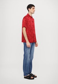 Rode shirt met korte mouwen met een gestructureerd bloemenpatroon, gecombineerd met blauwe spijkerbroek en zwarte sandalen, weergegeven vanaf de zijkant.