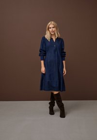 Hajszínű nő áll, sötétkék, térd alá érő, hosszú ujjú ruhát visel, fekete velúr térdig érő csizmával, barna háttér előtt.