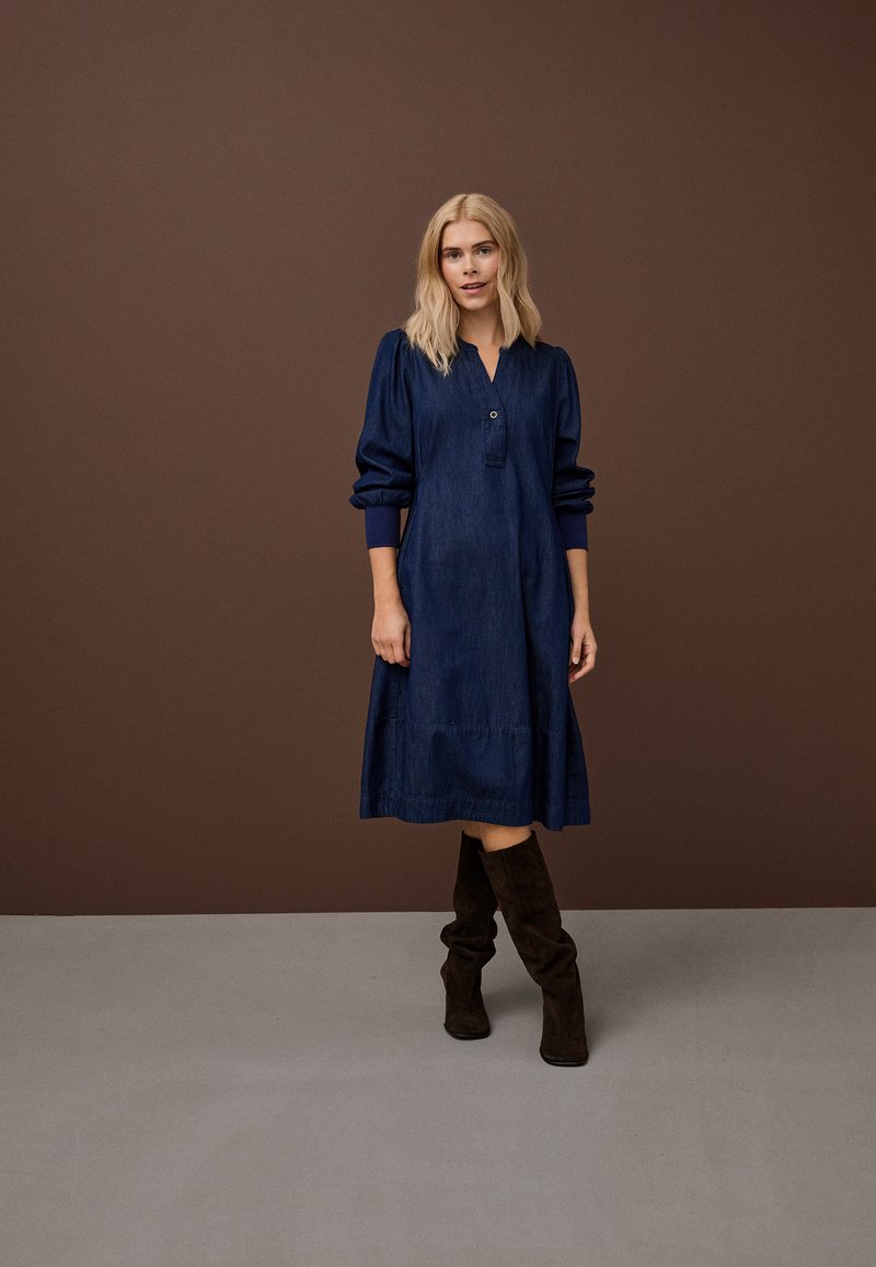 Hajszínű nő áll, sötétkék, térd alá érő, hosszú ujjú ruhát visel, fekete velúr térdig érő csizmával, barna háttér előtt.