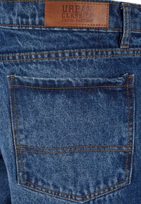 Blauwe spijkerbroeken van denim met een leren patch met de tekst "Urban Classics Denim Factory." Heeft een achterzak met zichtbare stiksels.