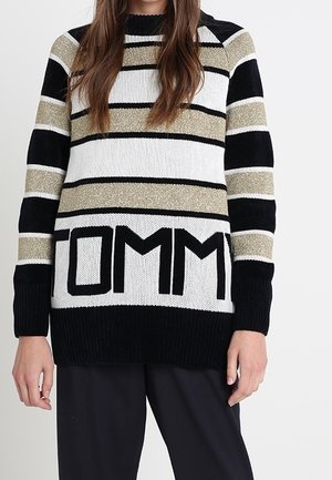 Femme portant un pull en maille rayé avec des bandes noires, blanches et dorées, et le mot "TOMMY" en grandes lettres noires sur le devant.