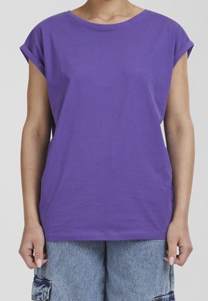 Person trägt ein schlichtes lila T-Shirt mit hochgekrempelten Ärmeln und hellblaue Denim-Cargoshorts, steht vor einem hellen Hintergrund.