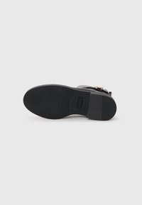 Semelle de chaussure noire avec surface en caoutchouc texturé, petit talon, et marquage de la marque. Présente une sangle et un accent en boucle en métal.