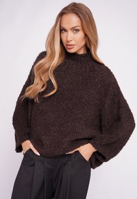 Brown textured sweater with a high neck and loose, oversized fit, paired with black trousers. Soft material with a cozy appearance.