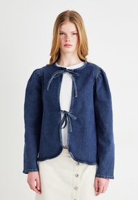 Denimjakke i mørkeblå med lange ermer, rund kant og lukking foran med knyting. Har et strukturert stoff og kontrastfargede sømmer.