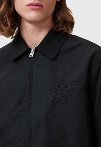 Čierna ľahká bunda s predným zipsom, bodovým golierom a vyšitým logom v kruhovom dizajne na ľavej časti hrudníka.