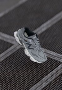 Grau- und schwarze New Balance Sneaker mit weißer Sohle, die auf einer Metallgitteroberfläche mit kreuzenden Linien ruhen.