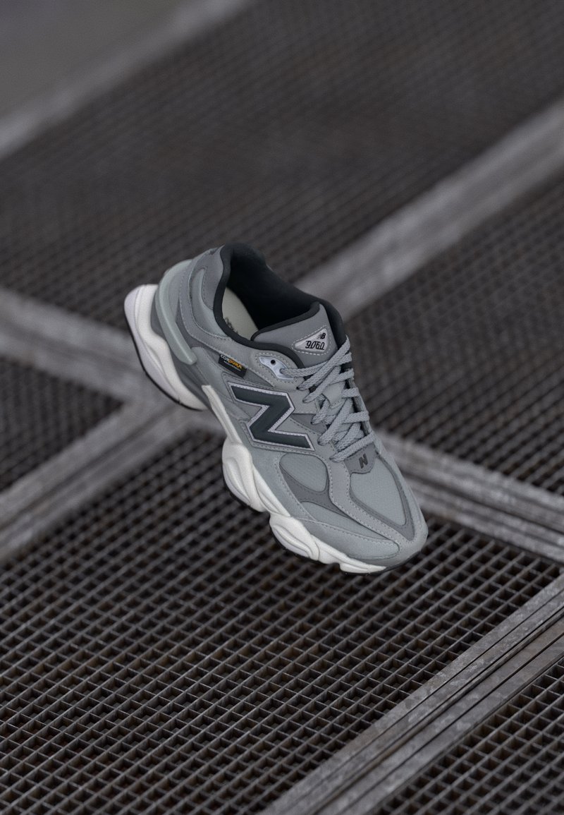 Zapatilla New Balance en gris y negro con suela blanca descansando sobre una superficie de rejilla metálica con líneas que se cruzan.