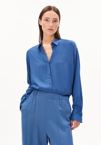 Blauwe satijnen blouse met een kraag, voorzien van een relaxte fit en knoopdetails. Gepaald met bijpassende blauwe broek met elastische tailleband.