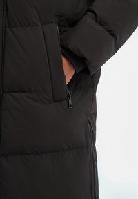 Schwarze Steppjacke mit Reißverschlusstasche, gesteppter Textur und schlanker Silhouette. Glatter Stoff mit matter Oberfläche.