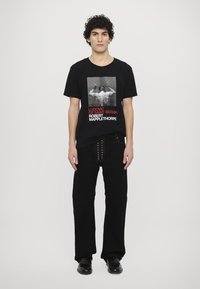 Schwarzes Baumwoll-T-Shirt mit einem grafischen Druck in einem rechteckigen Rahmen, mit fettem roten Text. Kombiniert mit schwarzen Hosen und Schuhen.