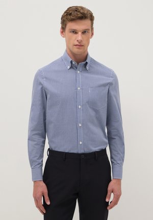 Hombre con camisa de cuadros azul y blanca metida dentro de pantalones negros, de pie contra un fondo liso.