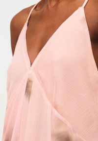 Κοντινό πλάνο ενός ατόμου που φοράει ροζ ανοιχτόχρωμο ρούχο χωρίς μανίκια, με V-λαιμόκοψη, λεπτές τιράντες και υφή υφάσματος.