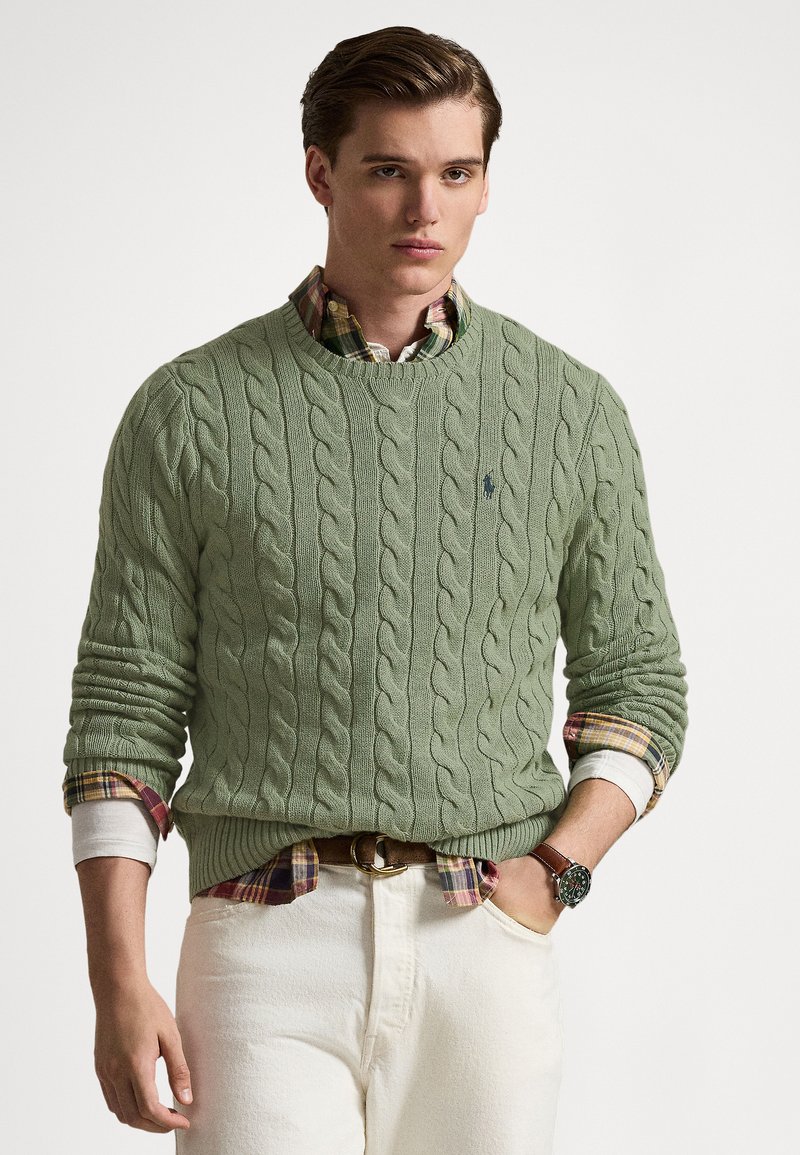 Cable-knit sweater in soft green, featuring a crew neckline and ribbed cuffs, layered over a plaid shirt, paired with white pants and a belt.