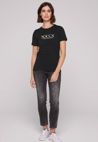 Camiseta negra con el texto blanco "SOCCX TIMLESS EDITION", combinada con jeans negros desgastados y zapatillas blancas. Diseño simple y ajustado.