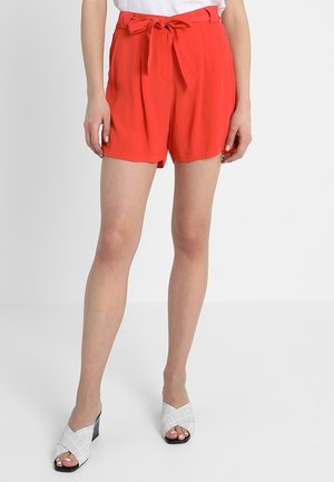 Rote, hoch taillierte Shorts mit einem Bindegürtel und gefaltetem Vorderteil. Getragen mit silbernen Absatzsandalen, die ein Kreuzdesign aufweisen.