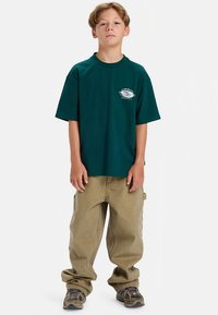 Donker petrolkleurig oversized T-shirt met een kleine witte afbeelding op de borst, gedragen met beige wijde broek en bruine sneakers met patroon.