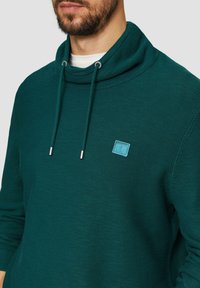 Turkusowa bluza z kapturem, o żeberkowej fakturze i srebrnych detalach. Zawiera małą, niebieską łatę z logo na piersi.