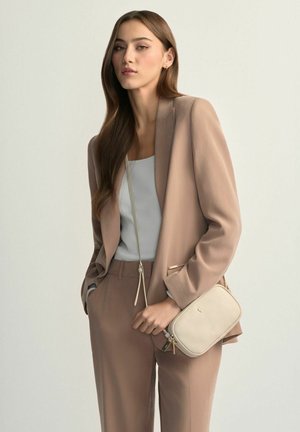 Kobieta w beżowym garniturze i białej bluzce trzymająca małą beżową torebkę na ramię, stojąca na tle jasnego, jednolitego tła.