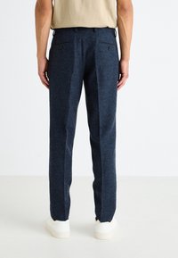Pantaloni blu navy in misto lana con una vestibilità sartoriale, dotati di due tasche posteriori e una texture liscia. Abbinati a sneaker bianche.