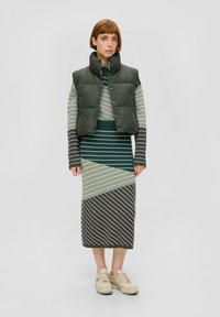 Puffige grüne Weste über einem gestreiften Strickkleid mit diagonalen Linien in Grün, Grau und Weiß; cremefarbene Sneaker runden das Outfit ab.