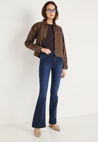 Chaqueta vaquera marrón con estampado de leopardo sobre una camiseta oscura, combinada con jeans acampanados azul oscuro y zapatos planos con punta afilada y textura.