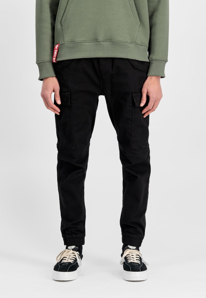 Personne portant un pantalon jogger cargo noir, des baskets noires et blanches, et un sweat à capuche vert avec une étiquette rouge sur le côté de la poche.