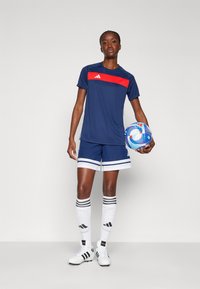 Uniforme deportivo azul marino con acento rojo, que incluye una camiseta de manga corta y pantalones cortos a juego, combinados con calcetines blancos y botas de fútbol.