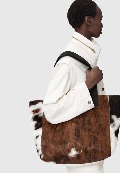 Personne en veste blanche portant un grand sac cabas en peau de vache marron et blanche avec des poignées noires sur l'épaule.