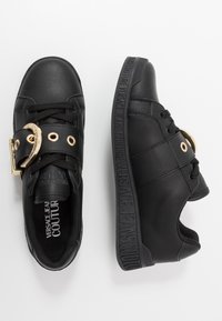 Baskets en cuir noir avec détails en métal doré, fermeture à lacets, panneaux latéraux texturés et logo embossé sur la semelle.