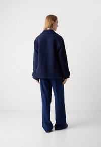 Chaqueta de mezcla de lana de color azul marino con un corte relajado, cuello ancho y detalle de abertura en la espalda, combinada con pantalones sueltos en azul marino.