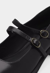 Czarne lakierowane buty Mary Jane z podwójnymi paskami i zapięciami na złote klamry, pokazane z bliska.