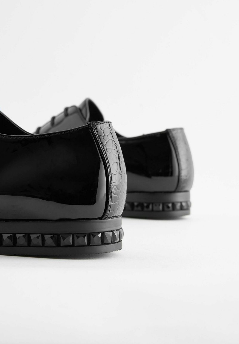 Ein Paar Schuhe aus schwarzem Lackleder mit texturiertem Absatz und Nietenbesatz an der Sohle, von hinten auf weißem Hintergrund gezeigt.
