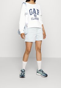 Sweatshirt blanc avec "GAP CLASSIC" imprimé en bleu foncé, shorts bleu clair, chaussettes blanches et baskets de sport bleu mixte. Sac à main bleu clair.